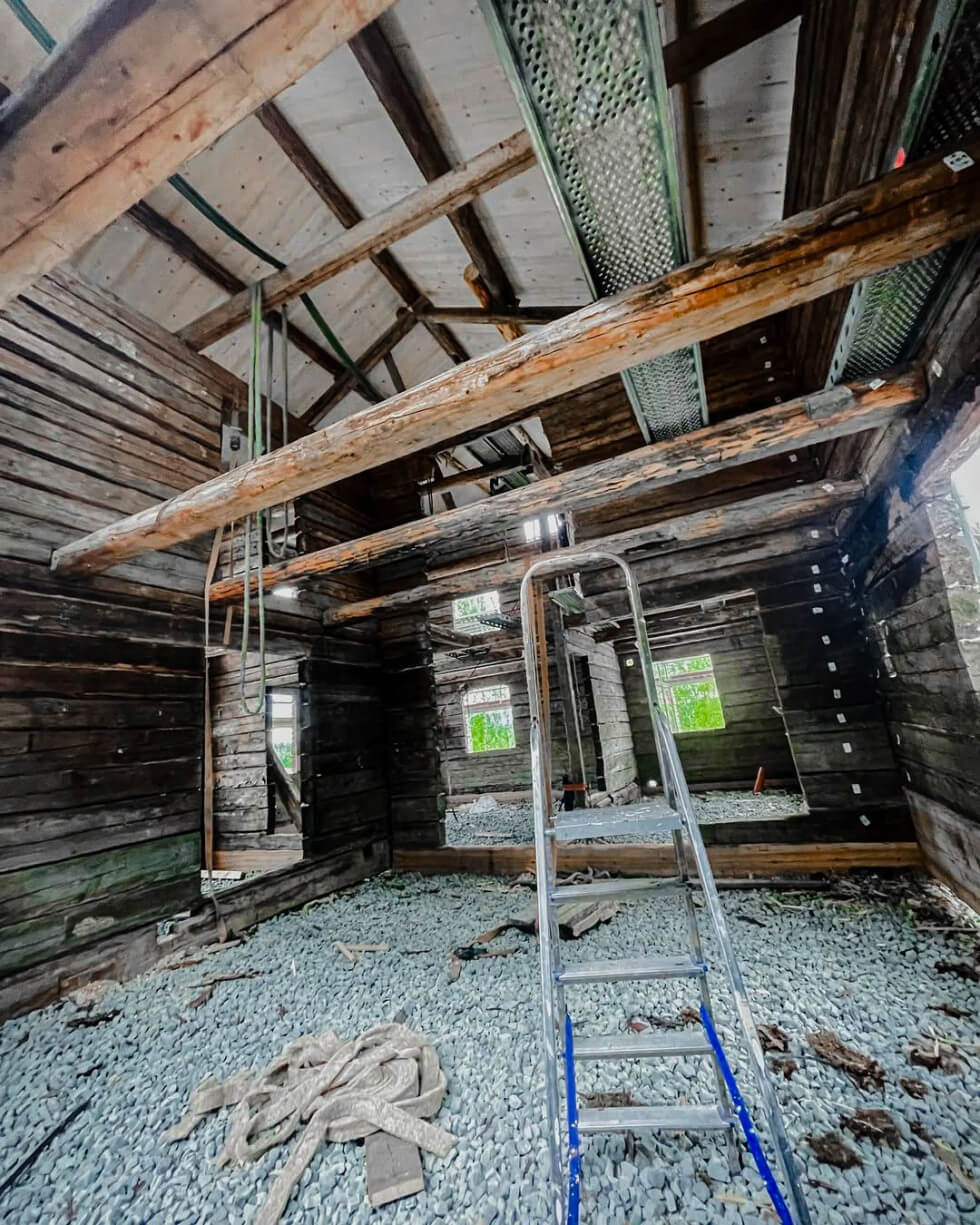 Interiör av en återuppbyggd timmerstomme med synliga stockväggar. Taket är täckt med ny råspont och byggställningar finns uppsatta. Golvet består av grovt grus och en stege står placerad i mitten av rummet.
