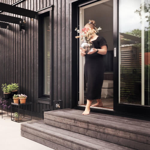 Kvinna går med vas i handen ut från sitt hus till altanen.